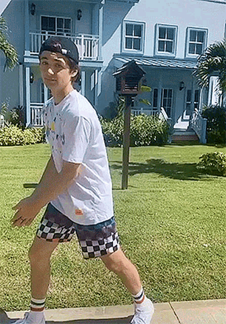 a young man in a white shirt and plaid shorts is walking in front of a house