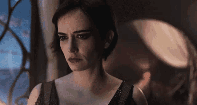 a woman in a black dress is standing in front of a window looking at the camera .