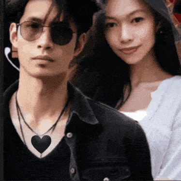 a man wearing sunglasses and a necklace with a heart on it stands next to a woman