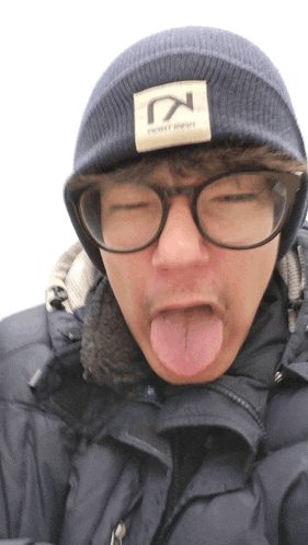 a man wearing glasses and a beanie with the letter k on it sticks his tongue out