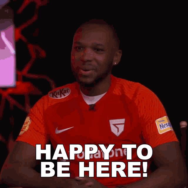 a man wearing a red kitkat shirt says " happy to be here "