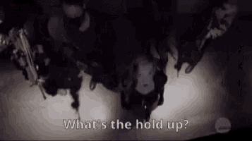 a group of people standing in a dark room with the words `` what 's the hold up '' .