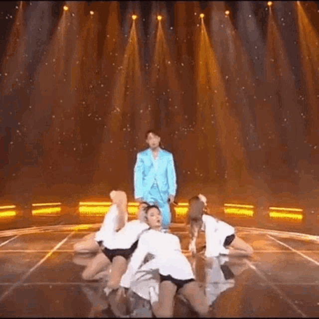 a man in a blue suit is standing on a stage surrounded by female dancers .
