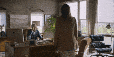 a man sits at a desk with an apple computer and a woman stands in front of him