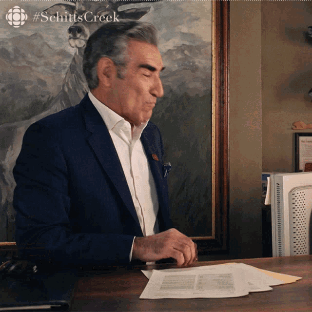 a man in a suit sits at a desk with #schittscreek written on the bottom
