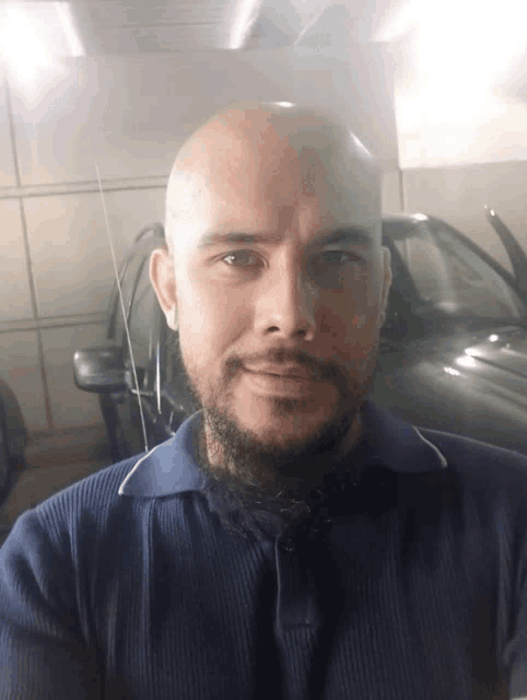 a bald man with a beard wearing a blue shirt stands in front of a car