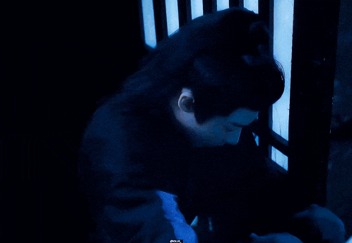 a man with long black hair is sitting in a dark room with a blue light behind him .