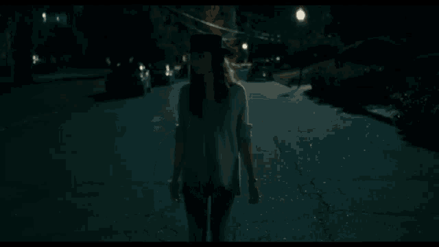 a woman is walking down a dark street at night wearing a hat .