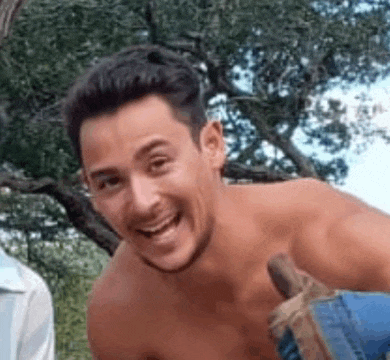 a shirtless man is smiling and looking at the camera while standing in front of a tree .