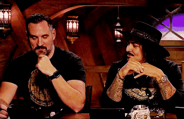 two men are sitting at a table with one wearing a hat and the other wearing a t-shirt with an eagle on it