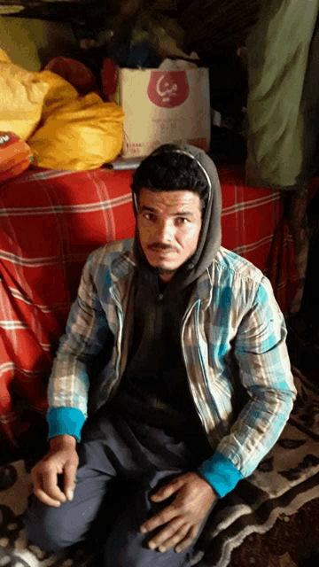 a man wearing a plaid shirt and a hooded jacket is sitting on the floor