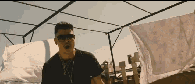 a man wearing sunglasses and a necklace stands in front of a clothes line