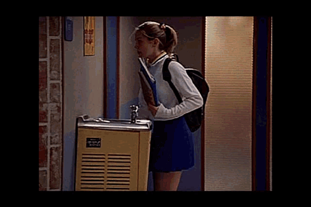 a girl in a cheerleader uniform is standing in front of a water fountain holding a book .