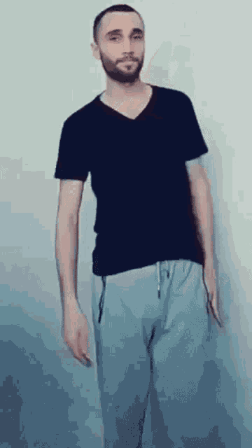 a man with a beard wearing a black shirt and grey pants is standing in front of a wall .