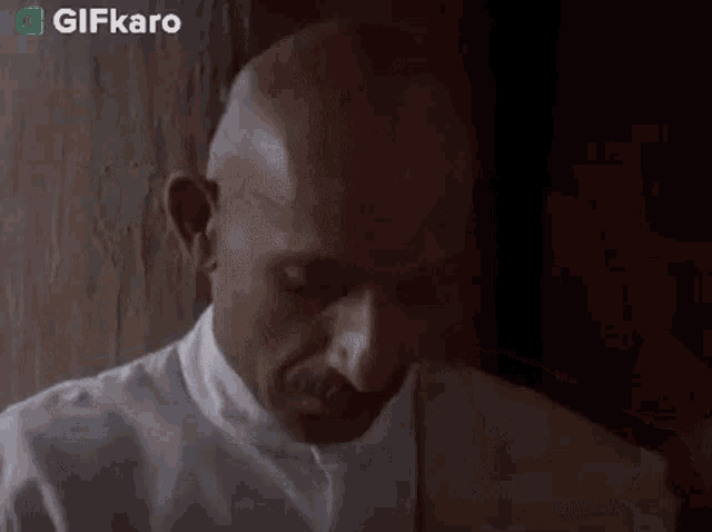a bald man with a mustache is leaning against a wall while holding a piece of paper in his hand .