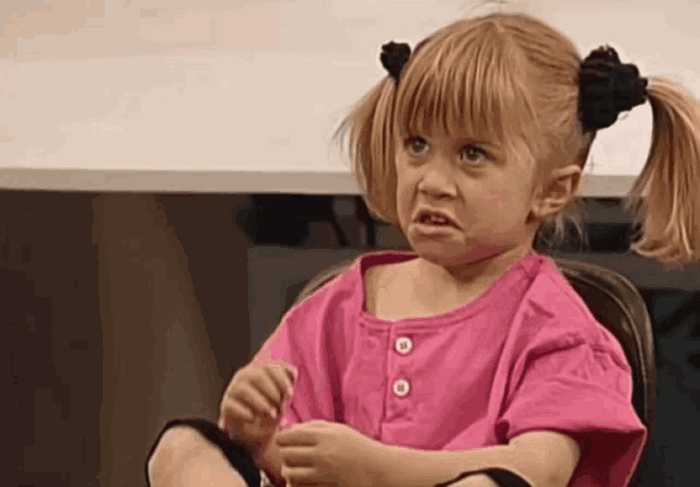 a little girl in a pink shirt is sitting in a chair making a funny face .