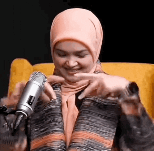 a woman in a hijab is sitting in a chair holding a microphone and smiling .