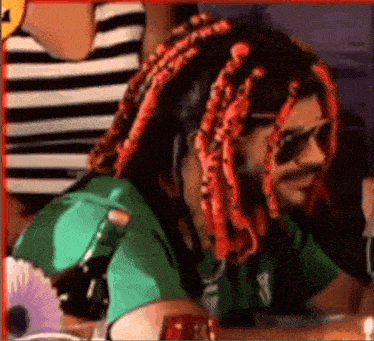 a man with dreadlocks and sunglasses is sitting at a table with a bottle of beer .