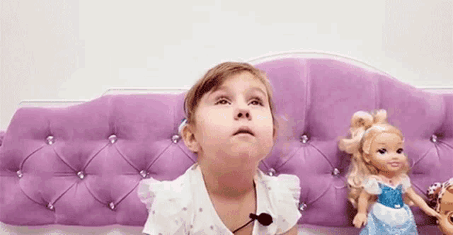 a little girl is sitting on a bed with a purple headboard and a doll .
