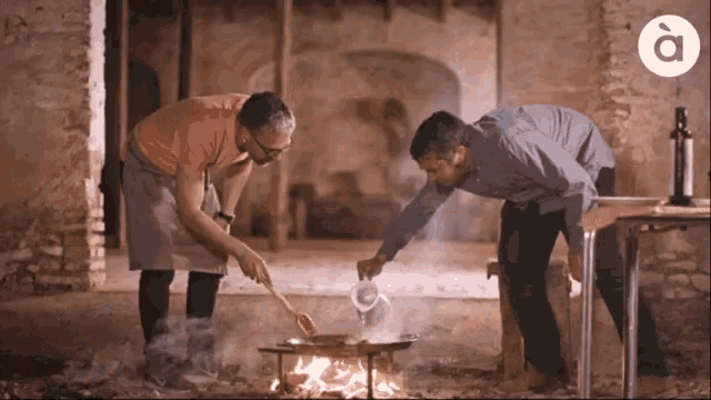 two men are cooking over a fire with the letter a on the bottom right