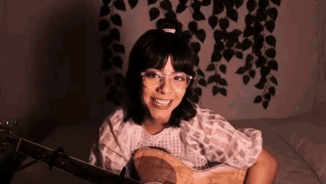 a woman wearing glasses is holding a guitar and smiling while sitting on a bed .
