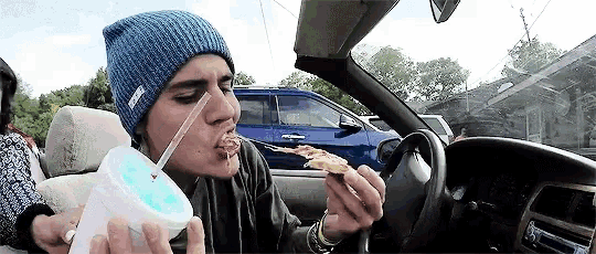 a man in a blue beanie is eating a slice of pizza in a car .