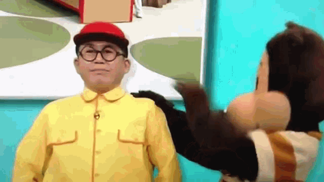 a man in a yellow suit and red hat is standing next to a stuffed animal .