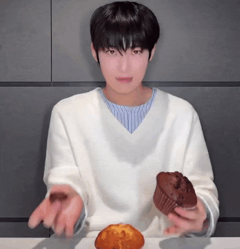 a young man in a white sweater is holding a chocolate muffin in his hands