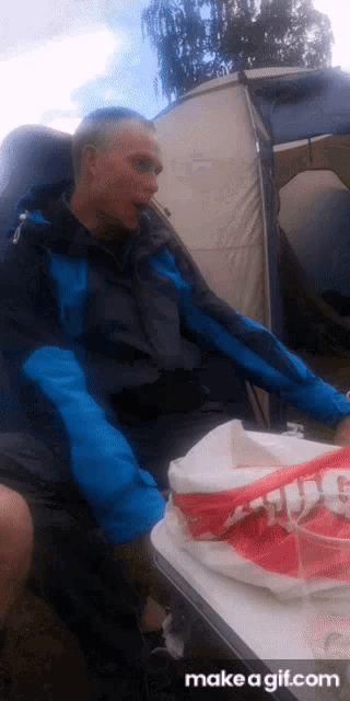 a man in a blue jacket sits in front of a tent with a bag that says ' chicken ' on it