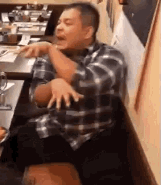 a man in a plaid shirt is sitting at a table in a restaurant with his hands outstretched .