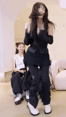 a woman in a black top and black pants is dancing while another woman sits on a chair .