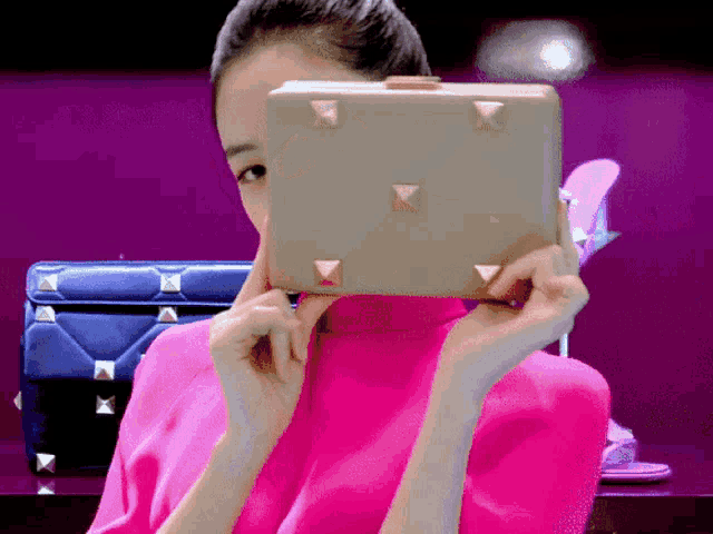 a woman in a pink top is covering her face with a purse with studs on it