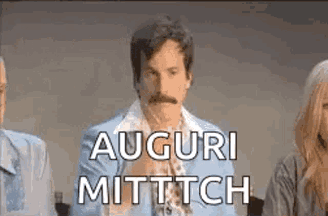 a man with a mustache is sitting in front of a group of people and says auguri mittch .