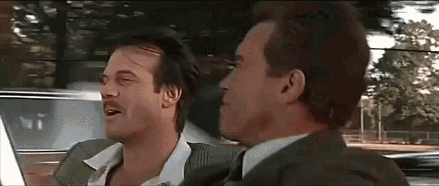 two men in suits and ties are sitting in a car and talking to each other .