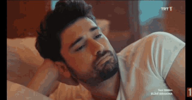 a man with a beard is laying on a bed with a trt 1 logo on the bottom