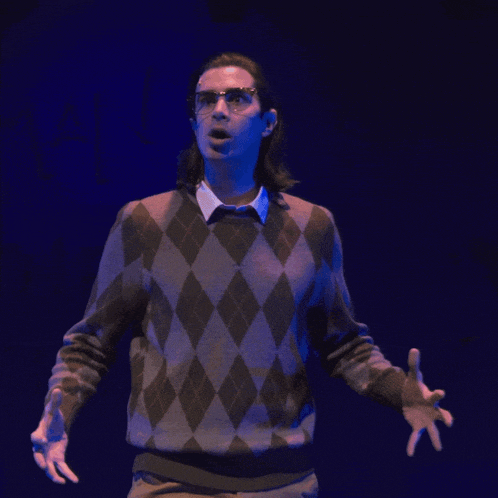 a man wearing glasses and a plaid sweater stands with his hands outstretched