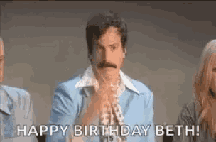 a man with a mustache is standing in front of a group of people and says `` happy birthday beth '' .