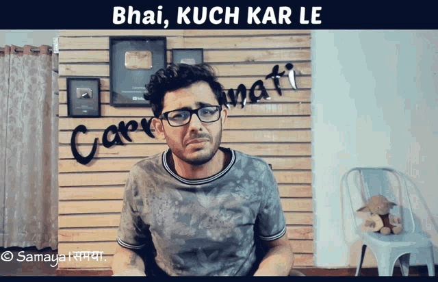 a man wearing glasses is sitting in front of a wall that says " bhai kuch kar le " on it