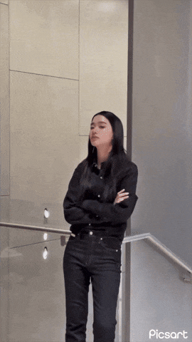 a woman in a black shirt and black jeans is standing in front of a wall with her arms crossed ..