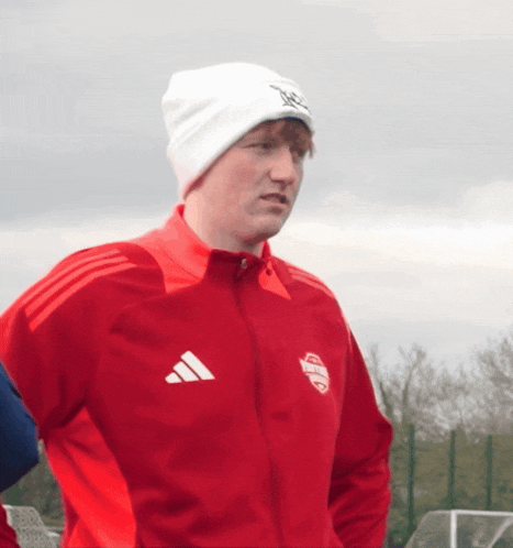 a man wearing a red adidas jacket and a white beanie stands in a field