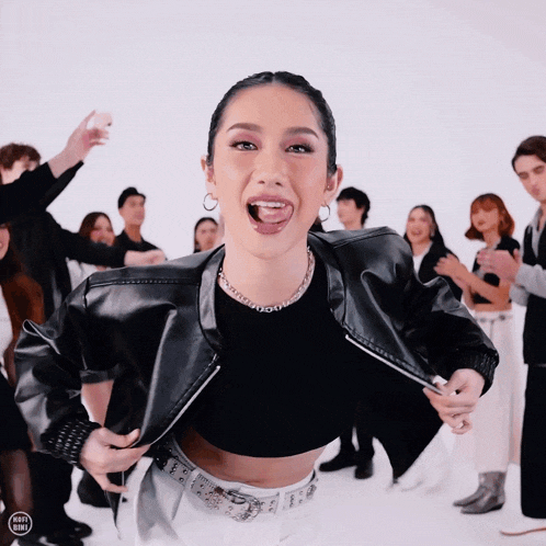 a woman wearing a black crop top and a leather jacket stands in front of a group of people
