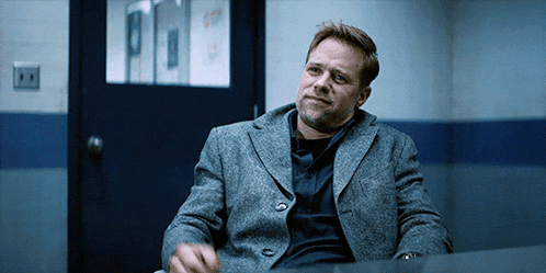 a man in a grey jacket sits at a desk in a police station