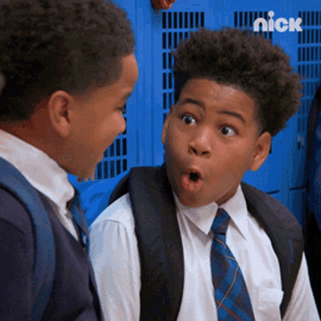 a boy with a surprised look on his face is talking to another boy in front of blue lockers that say nick