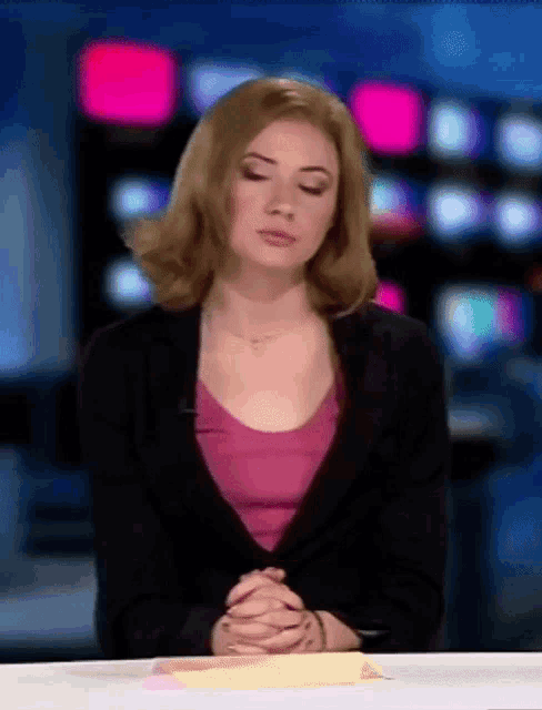 a woman in a pink shirt and black jacket sits at a desk with her eyes closed