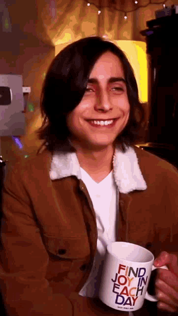 a young man in a brown jacket is holding a mug that says find joy in each day