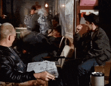 a man is reading a newspaper and smoking a cigarette while another man looks on