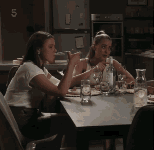 two women are sitting at a table with a bottle of water and a pitcher of lemonade