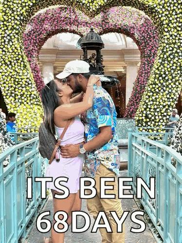 a man and a woman kissing in front of a heart shaped wall of flowers