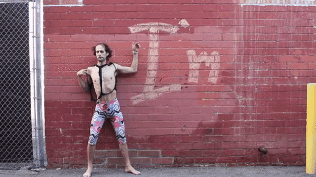 a shirtless man stands in front of a red brick wall with the letter t written on it