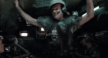 a man in a helmet and skull shirt is standing in a room with other people .